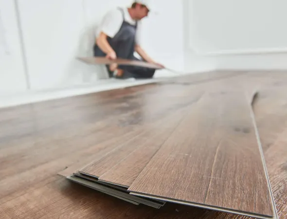A worker installs dark brown vinyl flooring planks in a room, with several planks stacked in the foreground and white walls in the background.